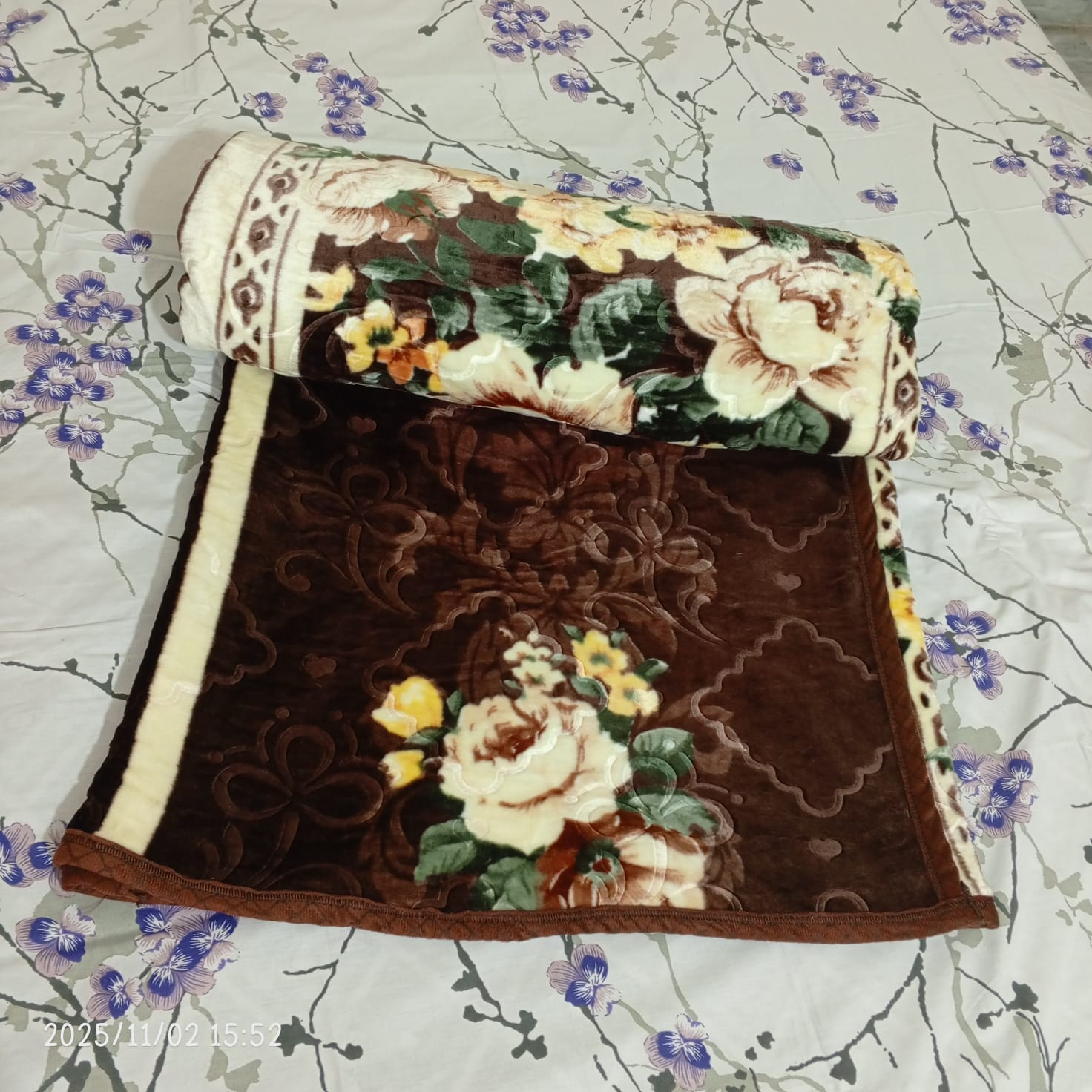 Brown floral blanket with cream and yellow rose design on a white bed sheet with purple flowers